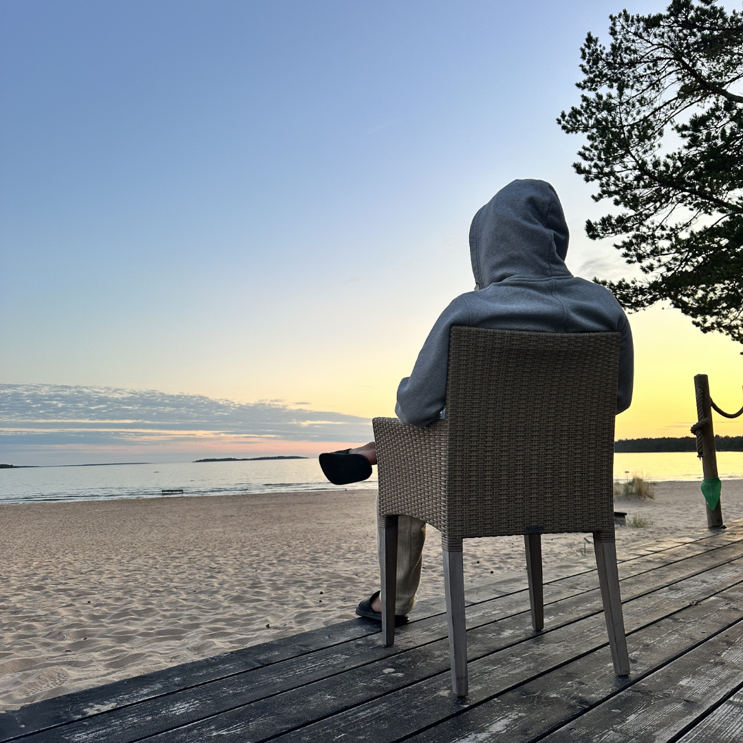 Harmaaseen huppariin pukeutunut henkilö istuu rottinkisessa tuolissa terassilla ja katselee tyyntä auringonlaskua rannalla.