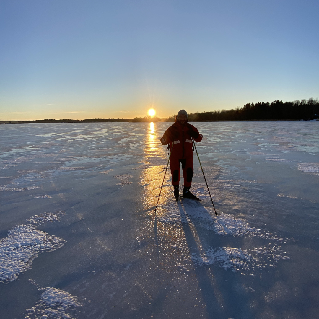 Henkilö luistelee jäätyneellä järvellä auringonlaskun aikaan, pitkät varjot ja talvinen puuraja kaukaisuudessa