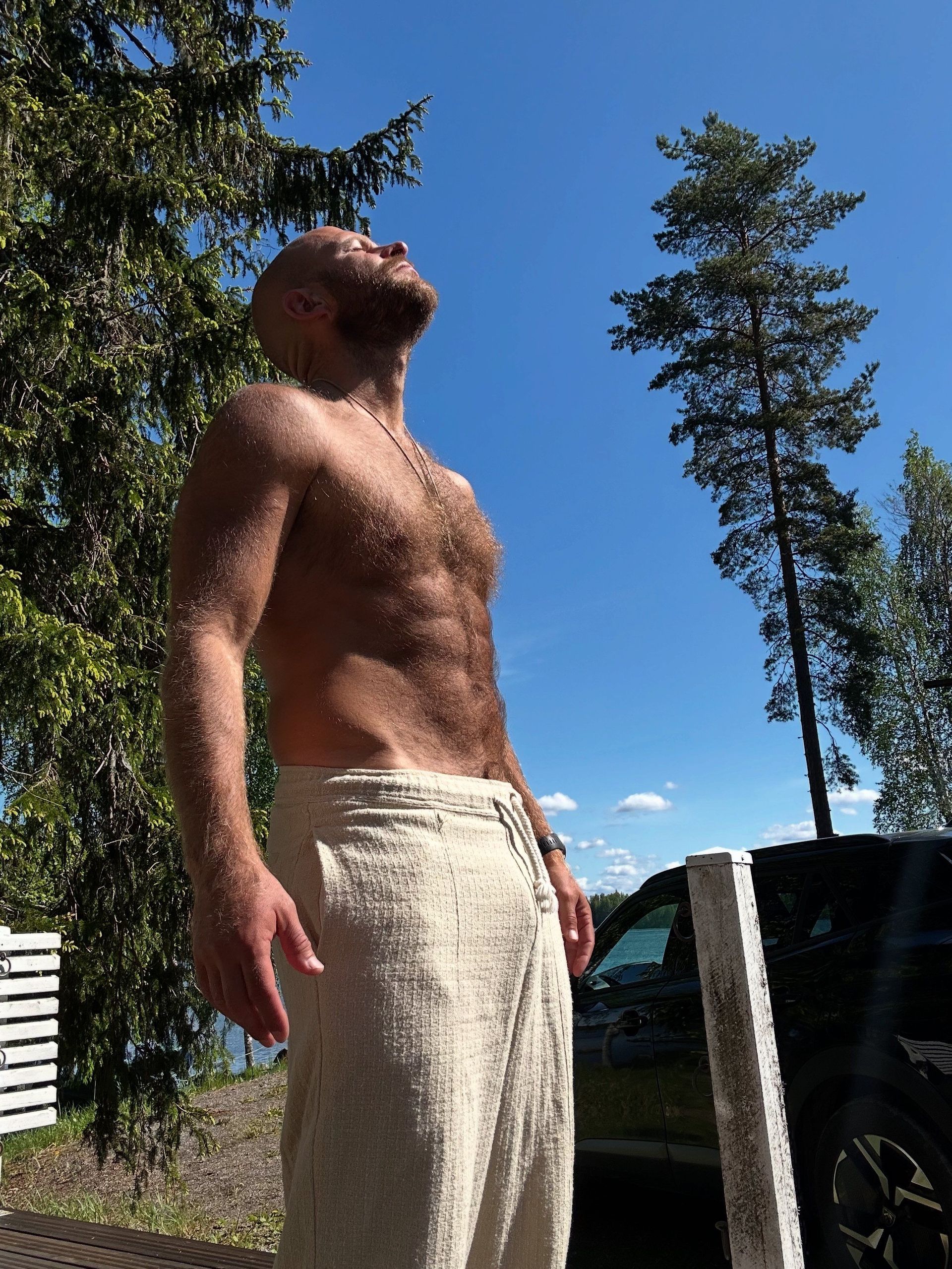 Paidaton mies seisoo ulkona kirkkaan sinisen taivaan alla puiden ja ajoneuvon vieressä, jalassaan kevyet housut.