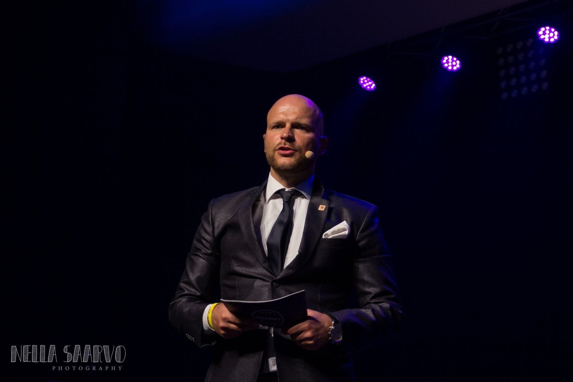 Man in a dark suit speaking on a dimly lit stage with purple spotlights