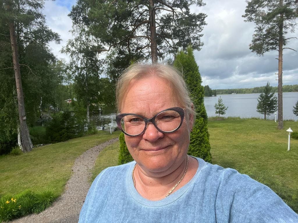 Nainen hymyilee ulkona järvenrantapolulla, silmälasit ja vaaleansininen paita päässään