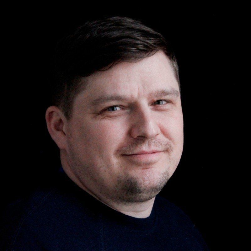 Muotokuva miehestä, jolla on lyhyet tummat hiukset ja tumma paita, ja joka katsoo kameraan mustaa taustaa vasten.