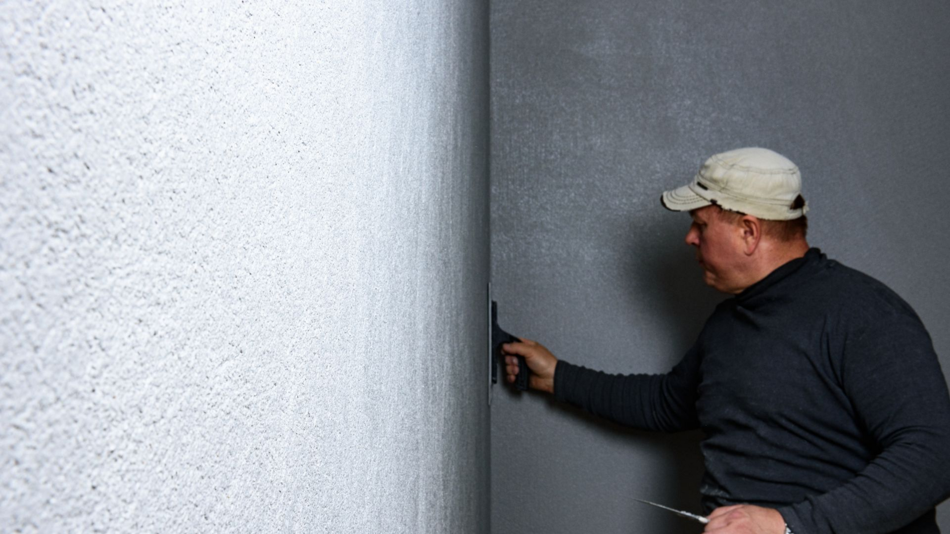 Un homme lisse le plâtre d'un mur à la truelle dans un coin ; les murs sont de couleurs différentes.