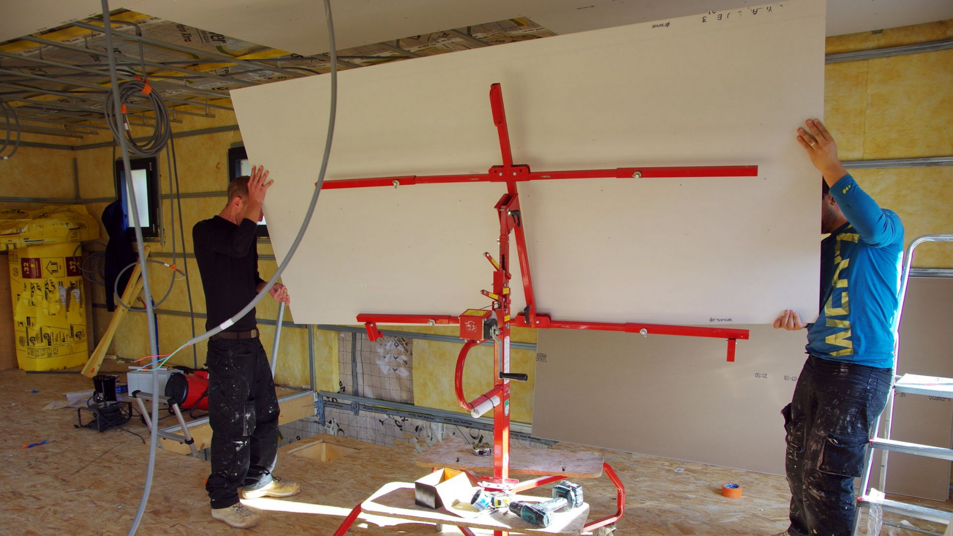 Deux ouvriers utilisent un élévateur à plaques de plâtre pour installer un grand panneau sur un plafond en construction.