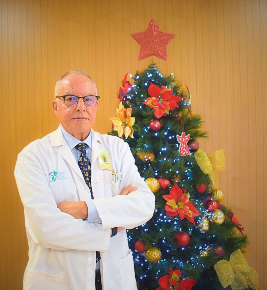 Un médico con bata blanca se encuentra junto a un árbol de Navidad decorado con una estrella roja en la cima.