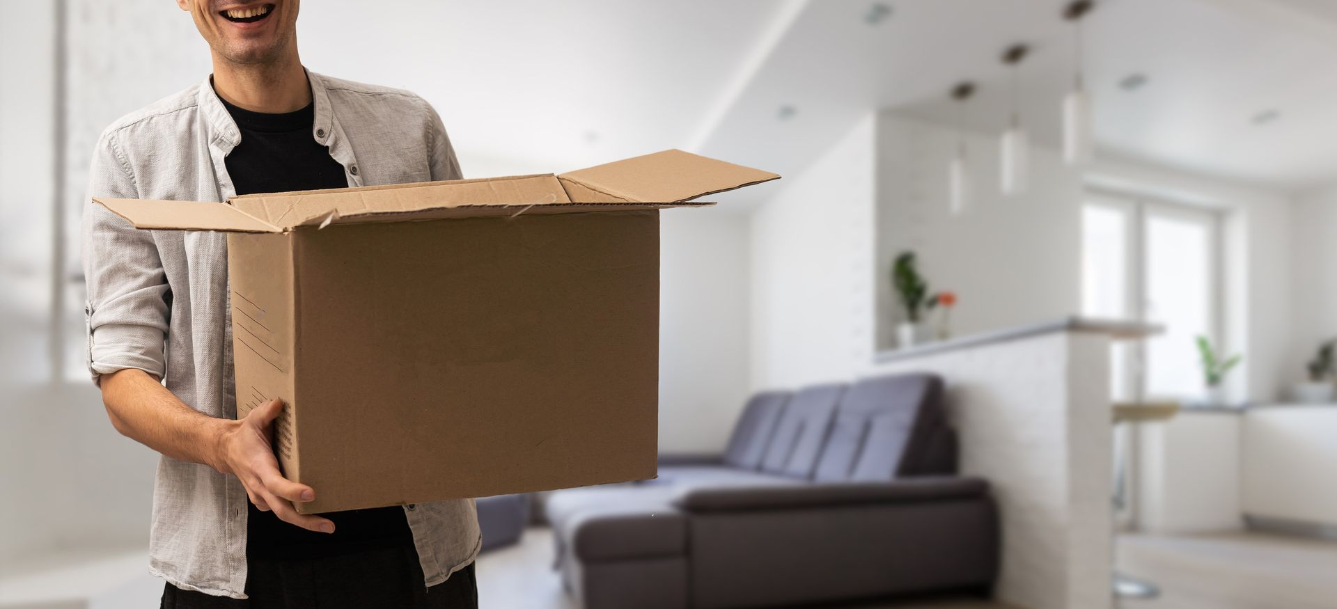 Una persona sonríe mientras sostiene una caja de cartón abierta, posiblemente mudándose a una casa.