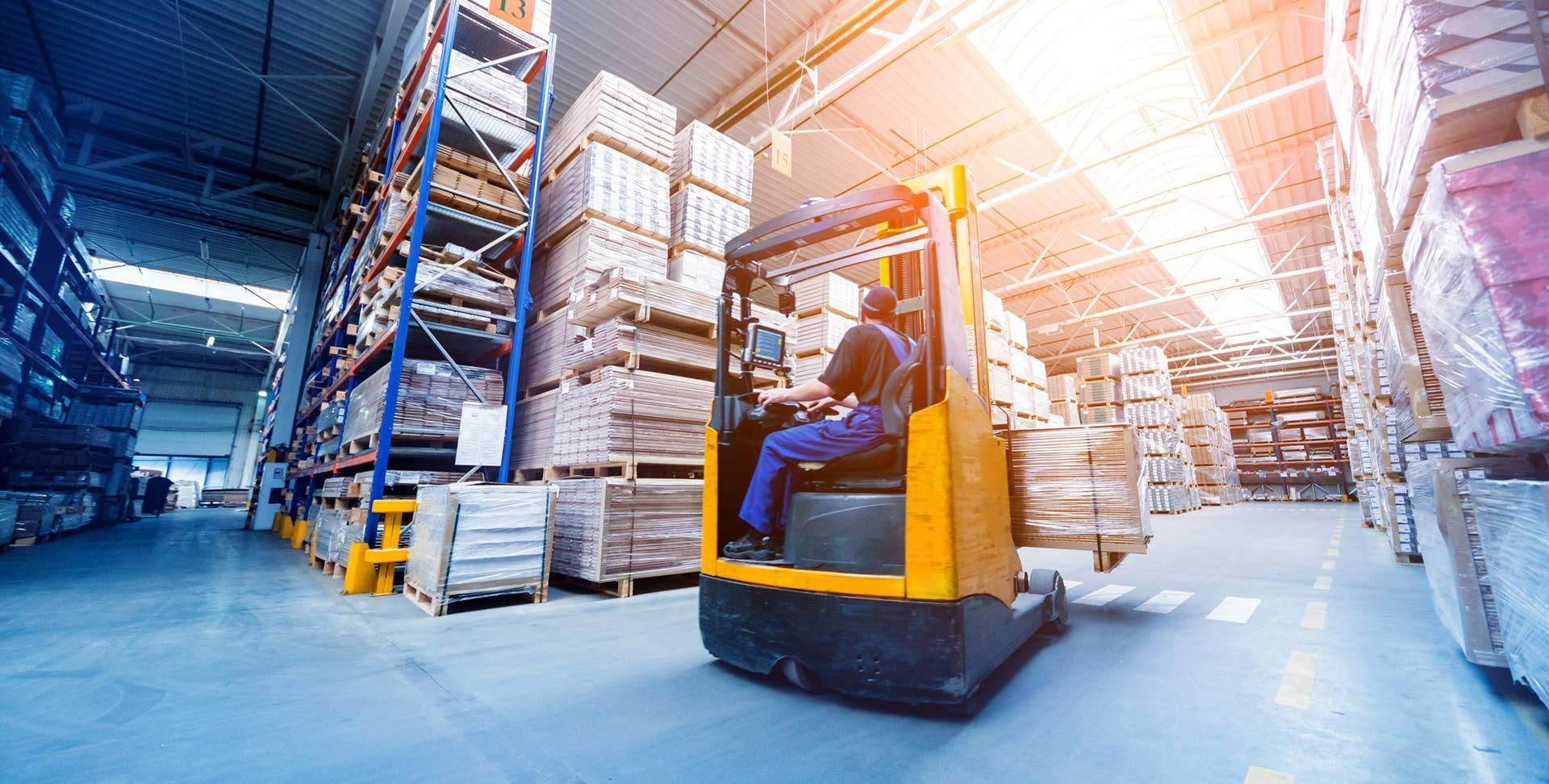 Operador de montacargas en un almacén, moviendo cajas entre estanterías altas. Luz brillante.