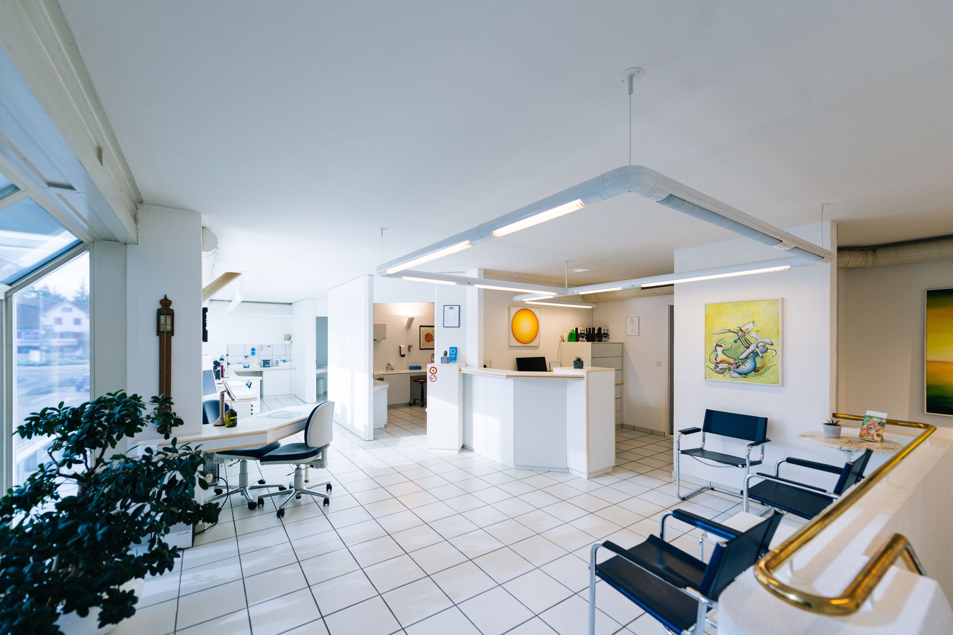 Strahlend weißer Empfangsbereich einer Arztpraxis mit Wartestühlen, Empfangstresen und Kunstwerken.