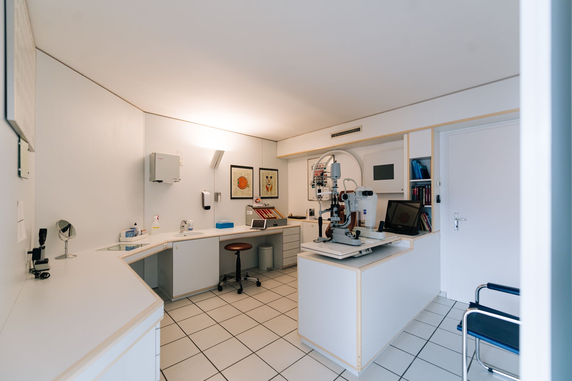 Ein Untersuchungszimmer eines Augenarztes mit weißen Wänden, Untersuchungsgeräten und einem Hocker.