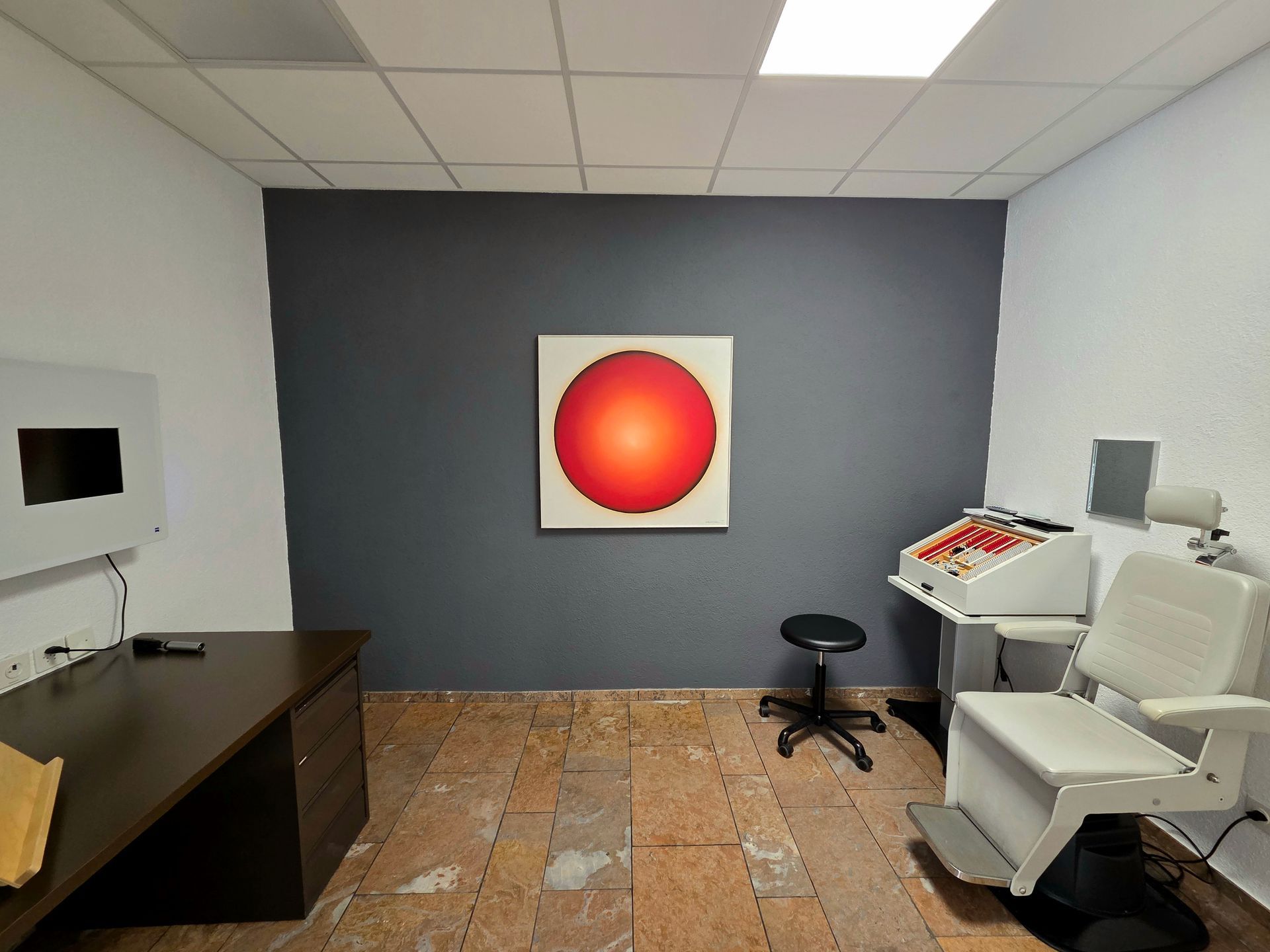 Eine Arztpraxis mit einem roten Kreis-Kunstwerk an einer dunkelgrauen Wand. Untersuchungsstuhl und Schreibtisch.