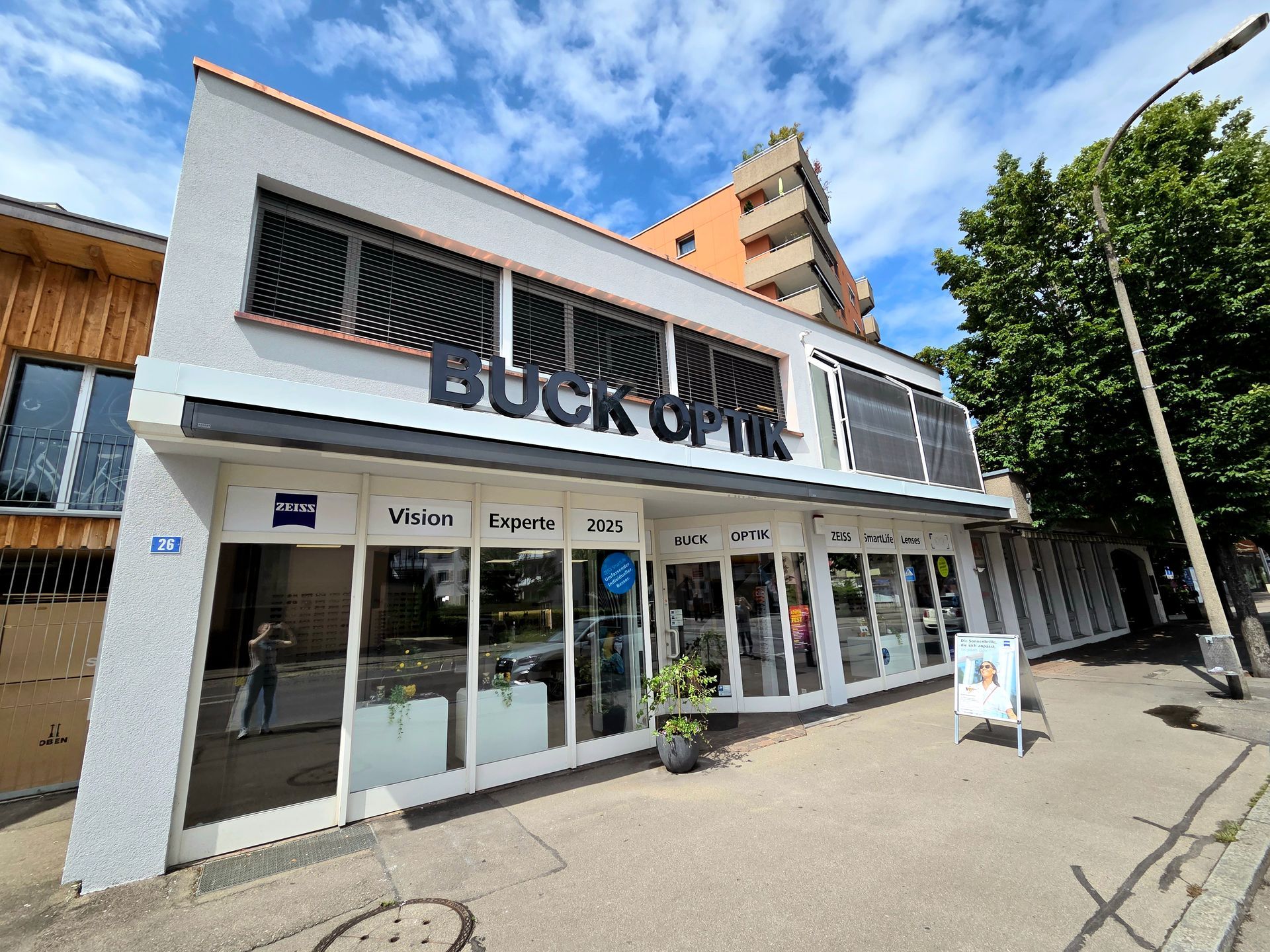 Schaufenster von Buck Optik mit weißer Fassade, schwarzer Schrift, Glasfenstern und blauem Himmel.