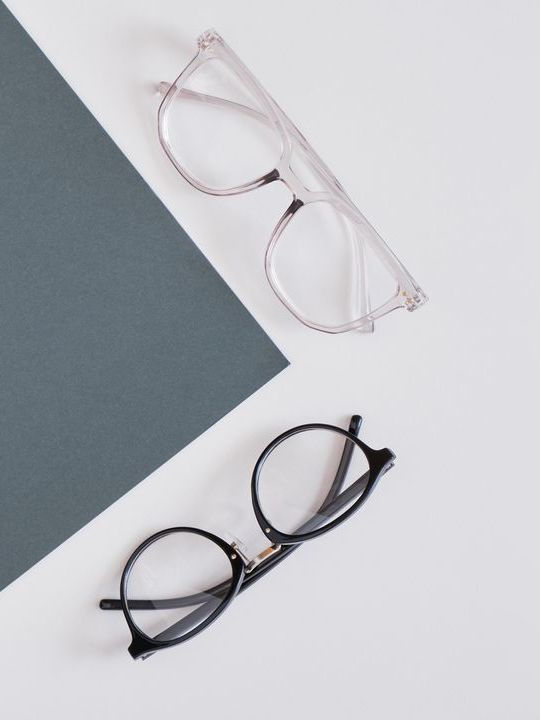 Zwei Brillen mit klarem und schwarzem Rahmen auf einer diagonalen Fläche in Weiß und Grau.