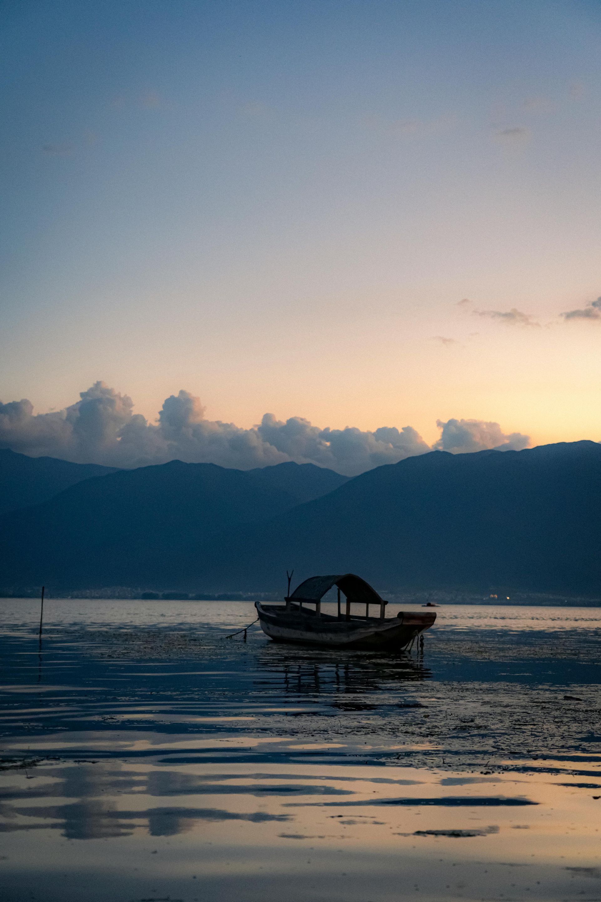 Boot auf einem See in der Abenddämmerung, Berge im Hintergrund. Das Wasser spiegelt die sanften Farben des Himmels wider.
