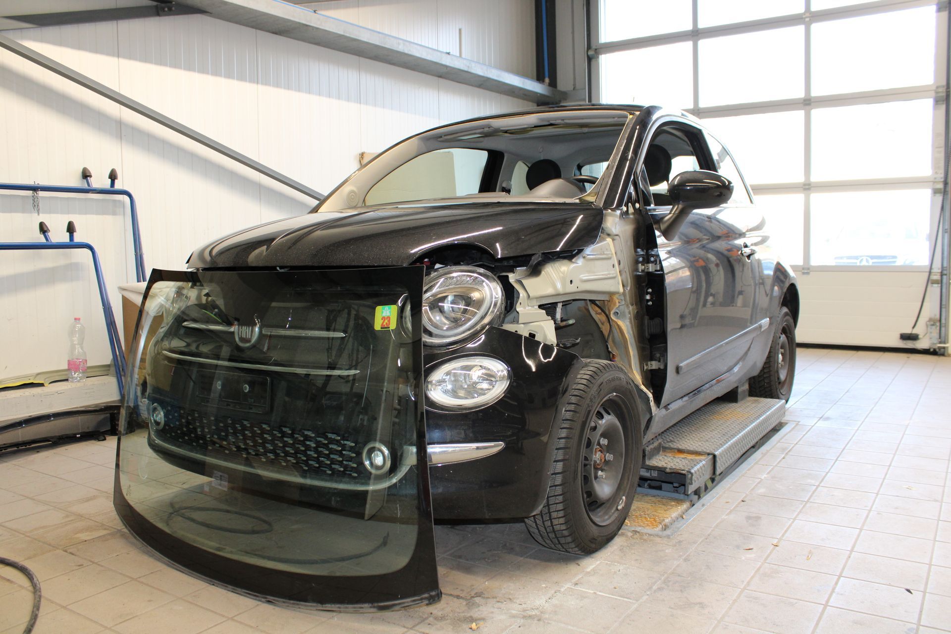 Ein schwarzes Auto mit kaputter Windschutzscheibe steht auf einer Hebebühne in einer Garage.