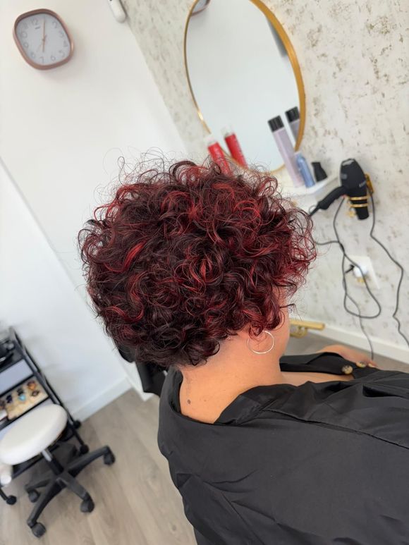 Mujer con cabello corto, rizado y con mechas rojas en una silla de salón; espejo y herramientas de peinado visibles en el fondo.