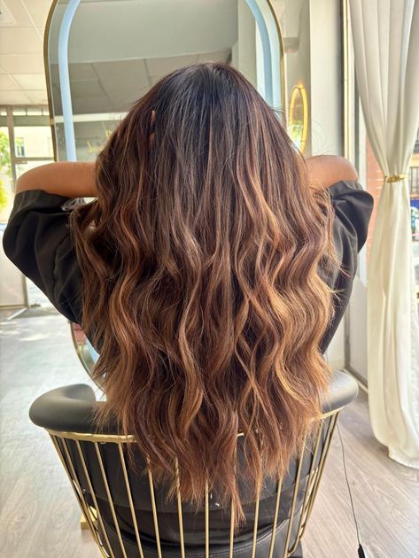 Mujer con cabello castaño largo y ondulado, estilo ombré. Sentada en una silla, con los brazos apoyados en el respaldo.