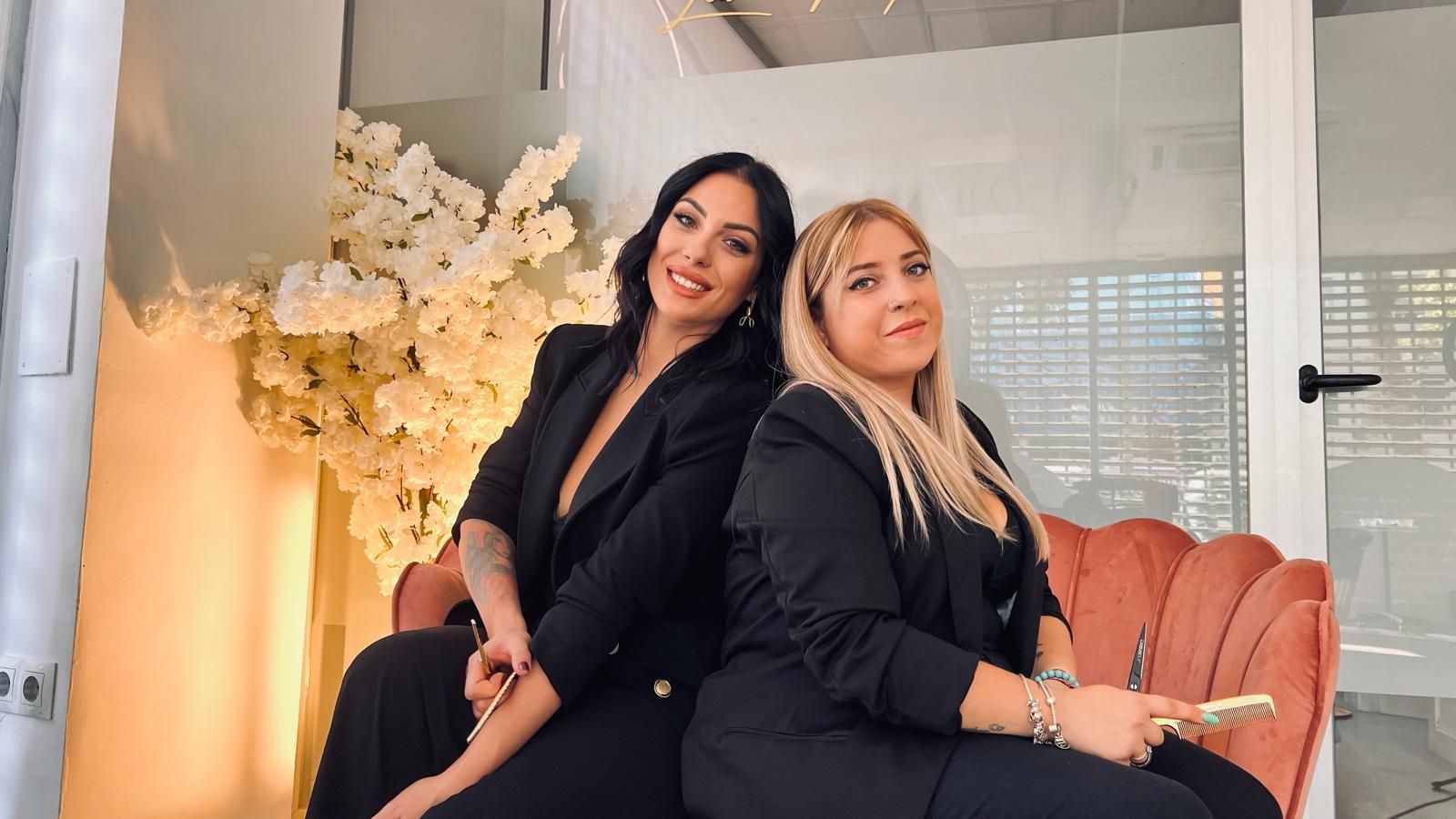 Dos mujeres con trajes negros sentadas espalda con espalda en un sofá rosa. Una sonríe, la otra mira a la cámara.