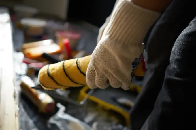 Una mano enguantada sostiene un rodillo amarillo sobre un banco de trabajo con herramientas y piezas metálicas.