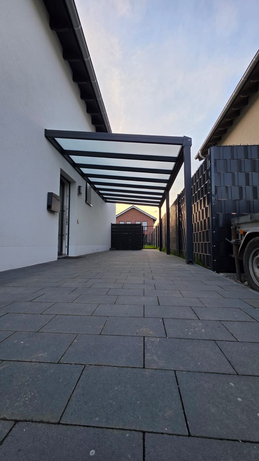 Eine moderne, dunkle Metall-Terrassenüberdachung ist an ein weißes Haus angebaut und erstreckt sich über eine grau gepflasterte Auffahrt.