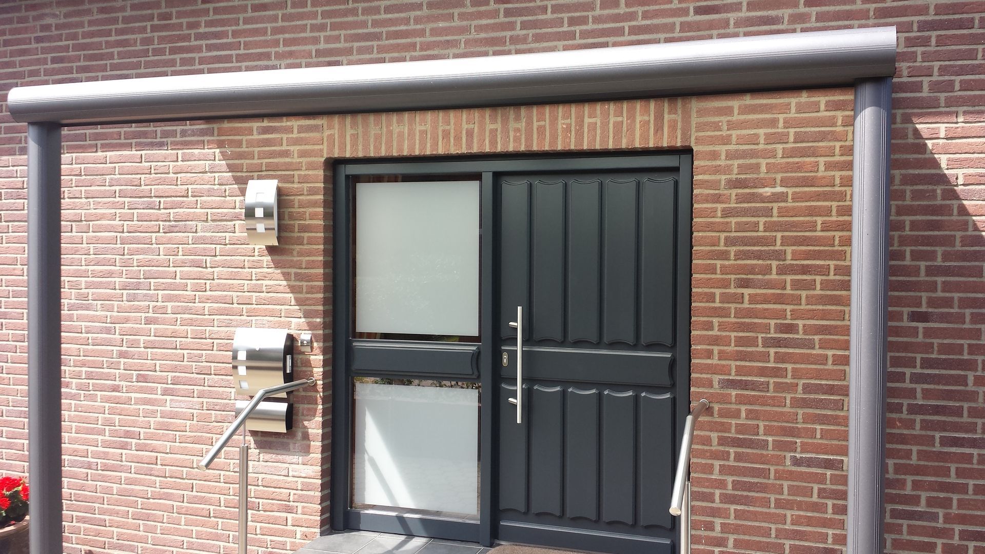 Eine moderne, dunkelgraue Eingangstür und ein Seitenfenster sind in eine rote Backsteinmauer eingelassen, unter einem überdachten Portikus mit metallenen Säulen.