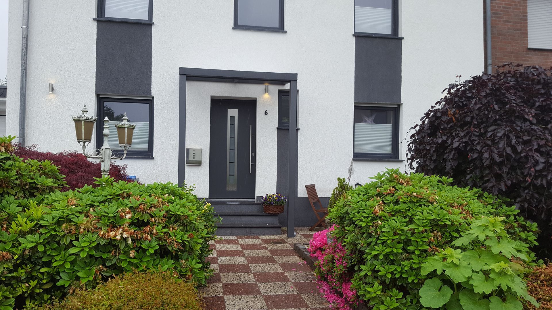 Moderner weißer Hauseingang mit dunkelgrauen Akzenten, passender Tür und üppigen grünen Büschen im Vorgarten.