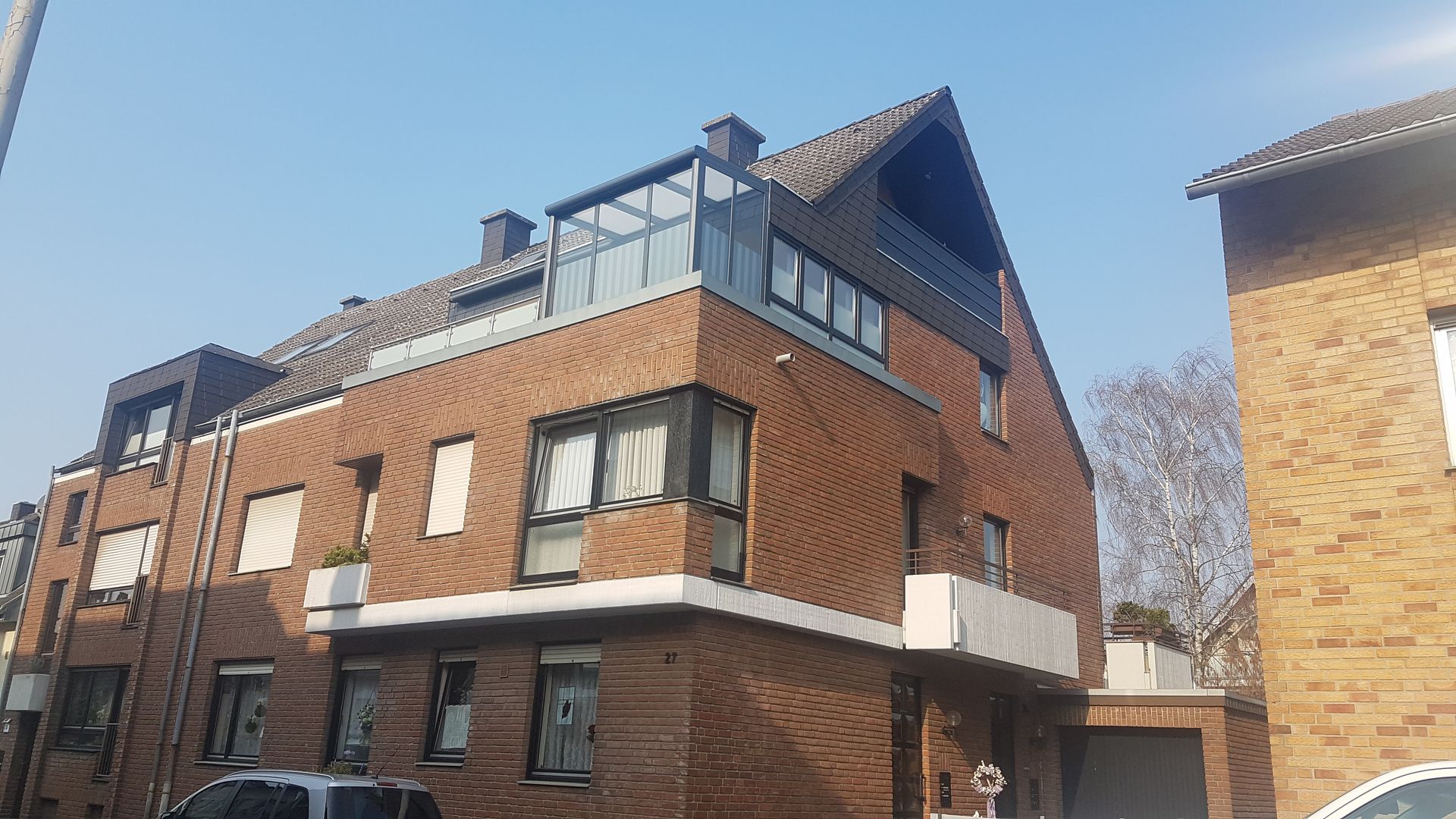 Ein mehrstöckiges Wohngebäude aus Backstein mit einer verglasten Dachterrasse unter strahlend blauem Himmel.
