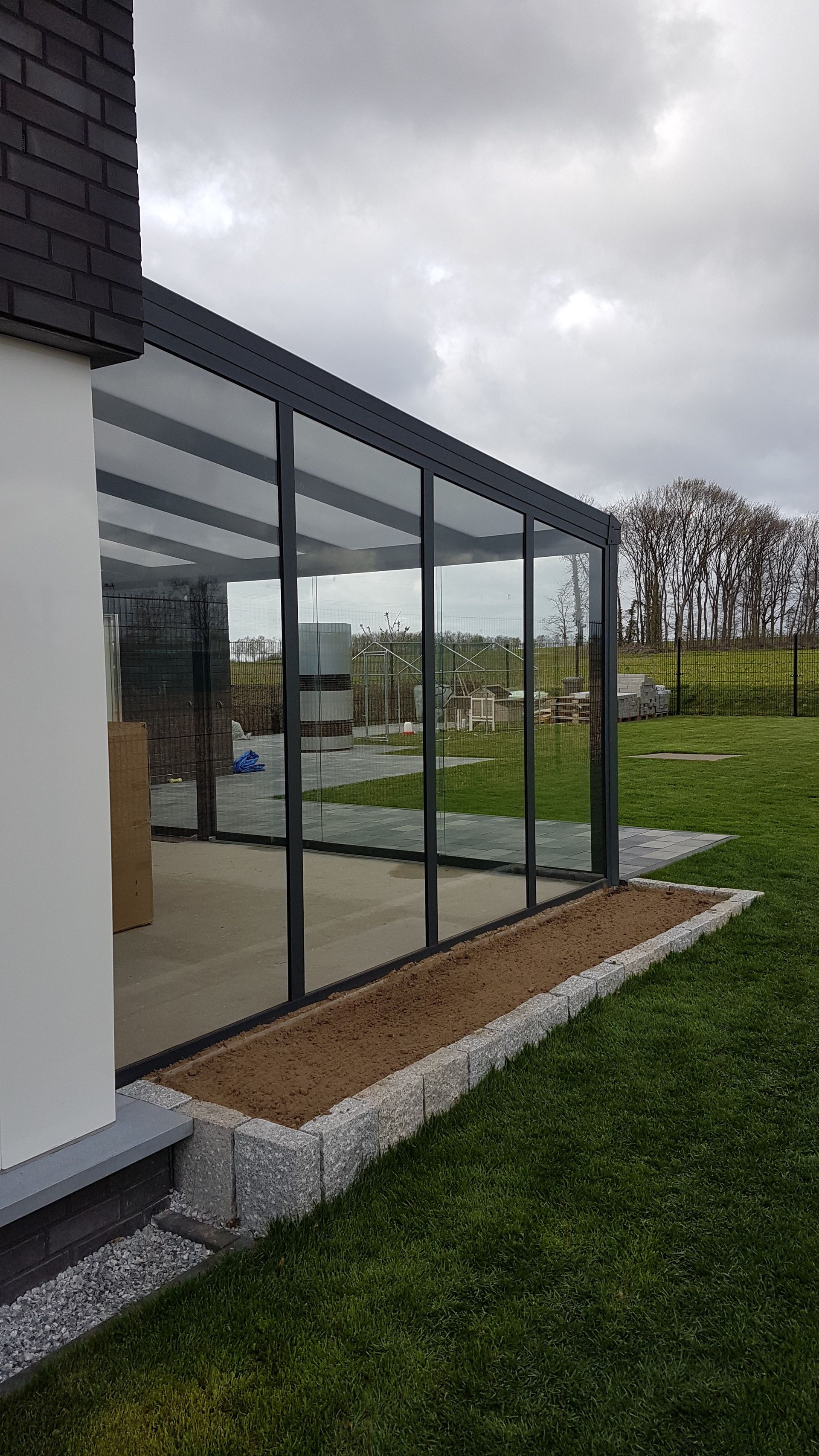 Eine moderne Terrassenüberdachung mit dunklem Metallrahmen und Glaspaneelen, die an ein Haus neben einem mit Steinen eingefassten Gartenbeet angebaut ist.