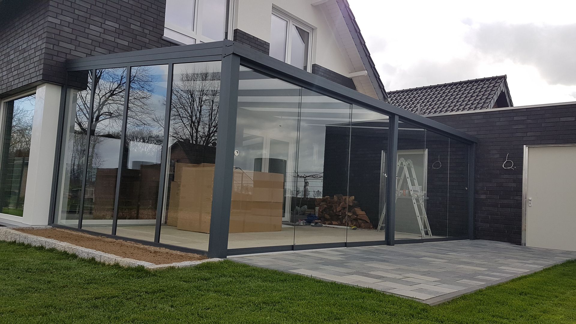 Eine moderne, dunkelgraue Terrassenüberdachung mit Glaswänden, die an ein Haus mit Steinfassade neben einem Rasen angebaut ist.