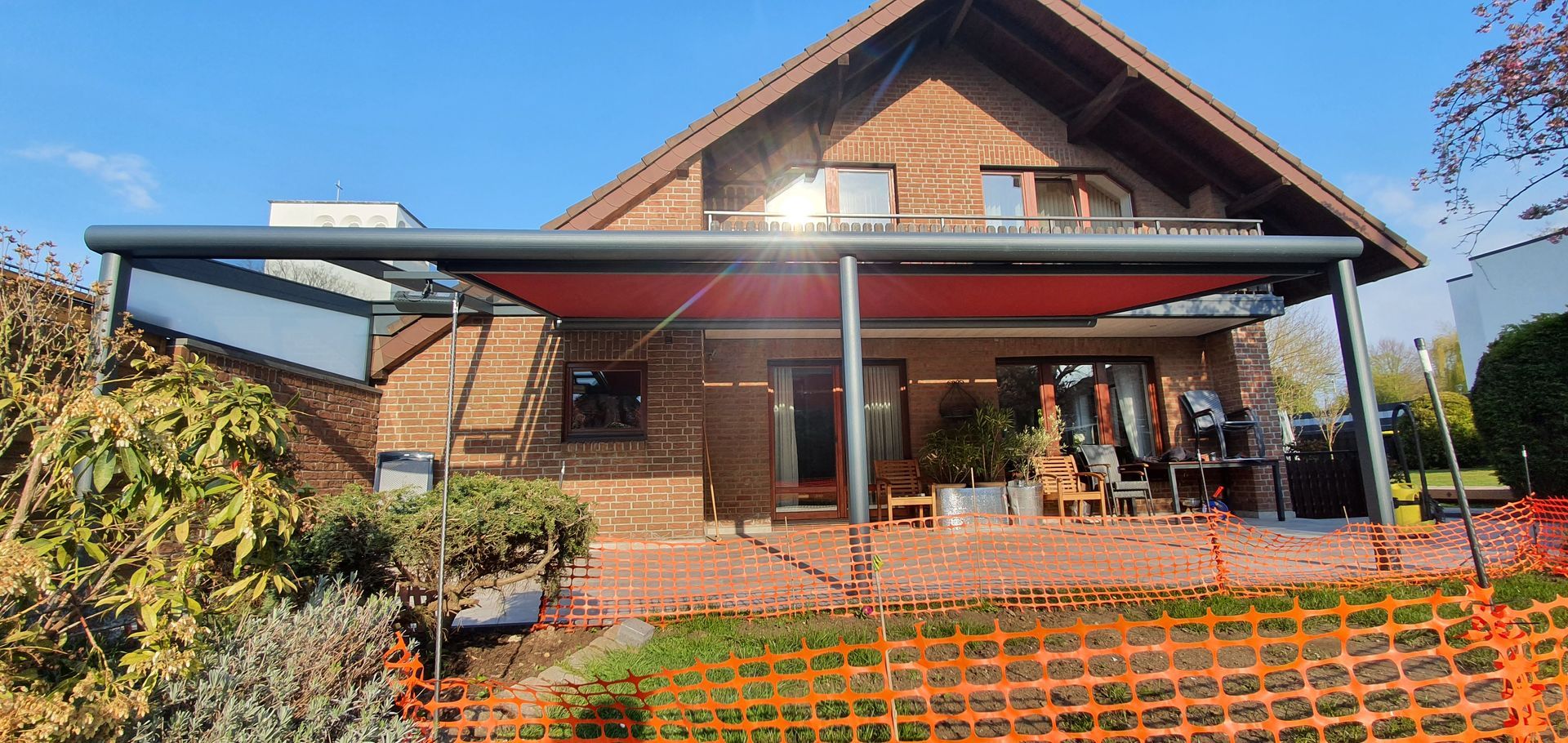 Ein Backsteinhaus mit einer großen, modernen dunkelgrauen Pergola, über der sich eine ausziehbare rote Markise über der Terrasse erstreckt.