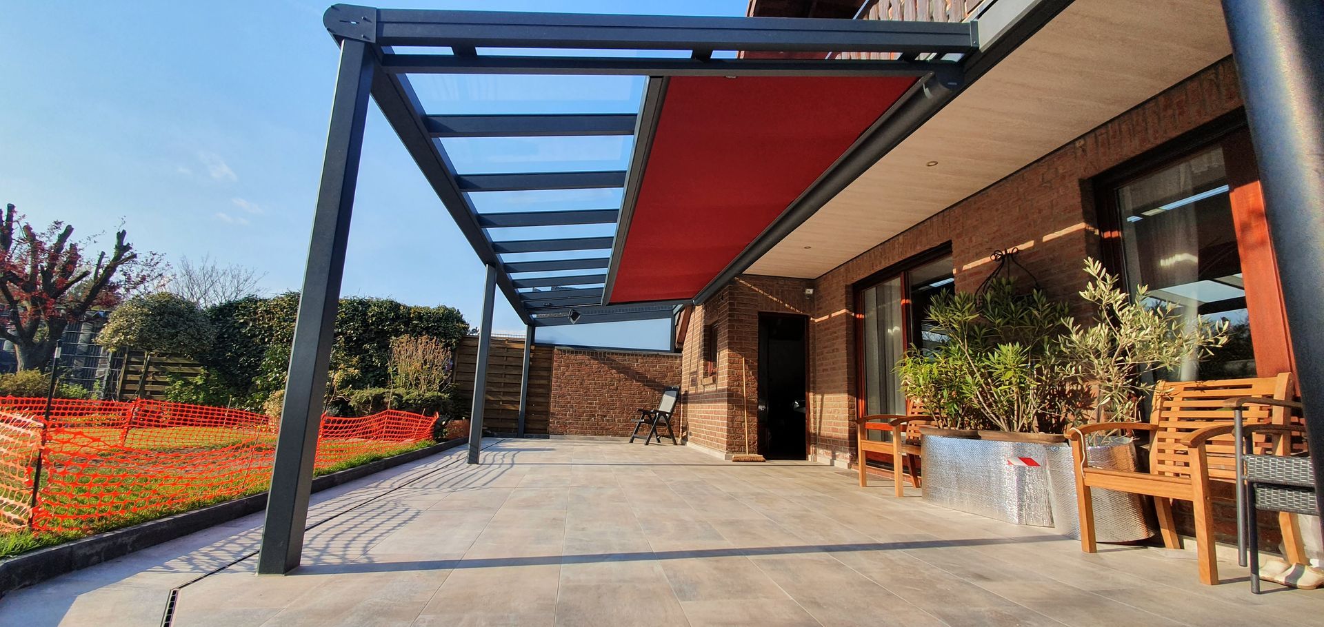 Eine Terrasse mit einer modernen Metallpergola mit einziehbarem rotem Vordach, Holzsitzgelegenheiten und einem großen Garten im Hintergrund.