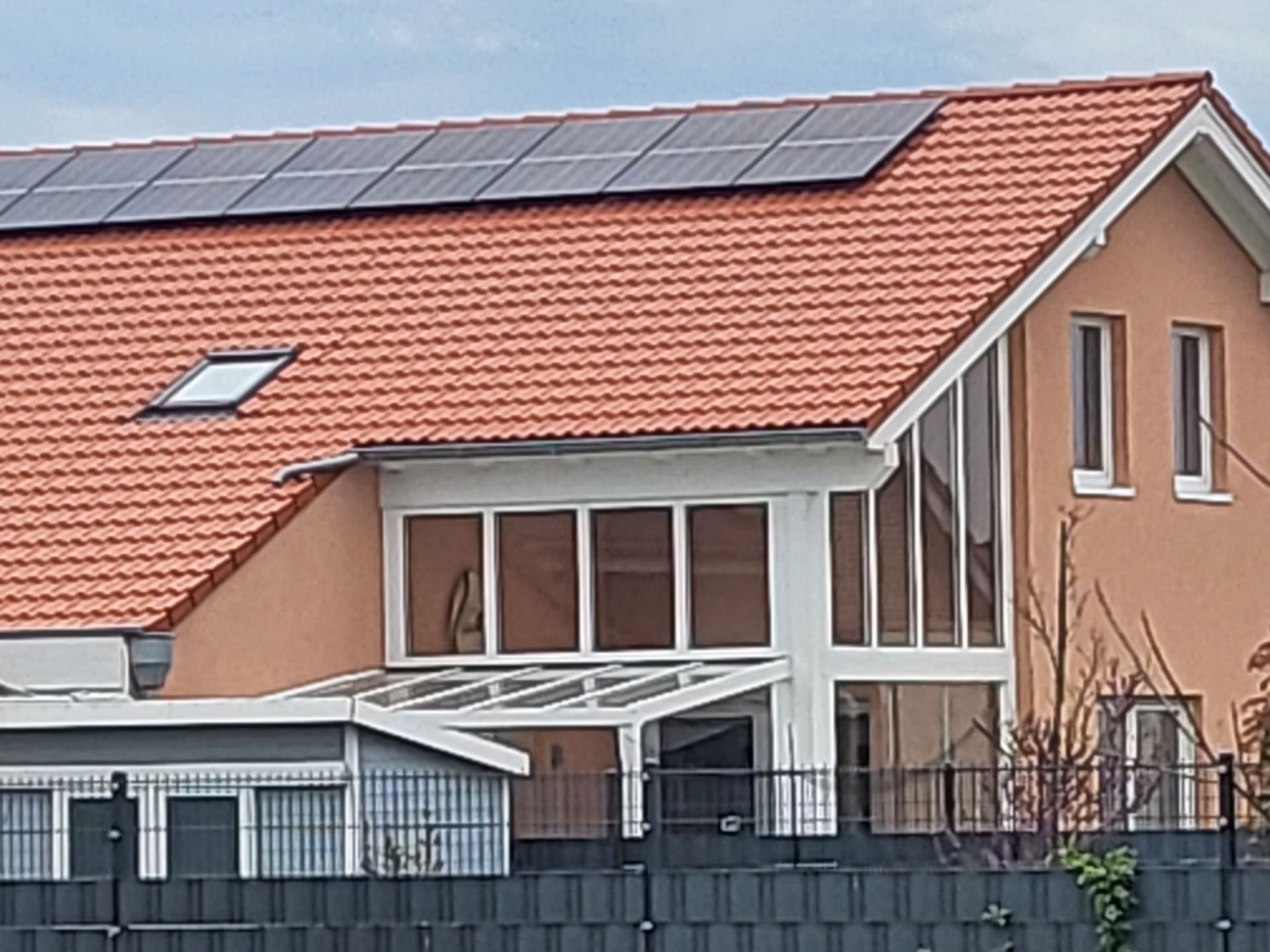 Ein modernes, pfirsichfarbenes Haus mit rotem Ziegeldach, Solaranlage, einem gläsernen Wintergarten und einem grauen Zaun.