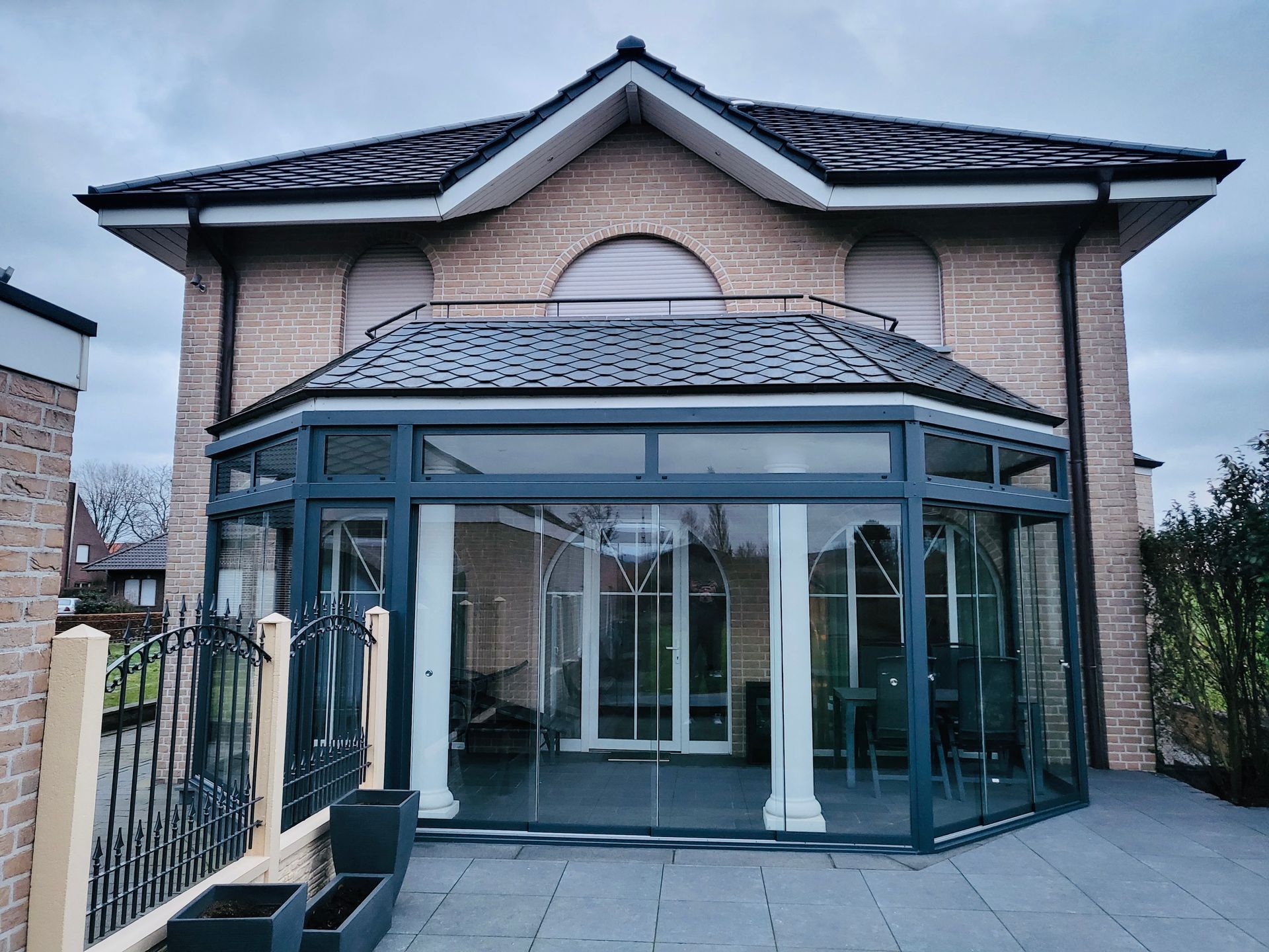 Ein Backsteinhaus verfügt über einen modernen, verglasten Wintergarten mit dunklem Rahmen auf einer Steinterrasse unter bedecktem Himmel.