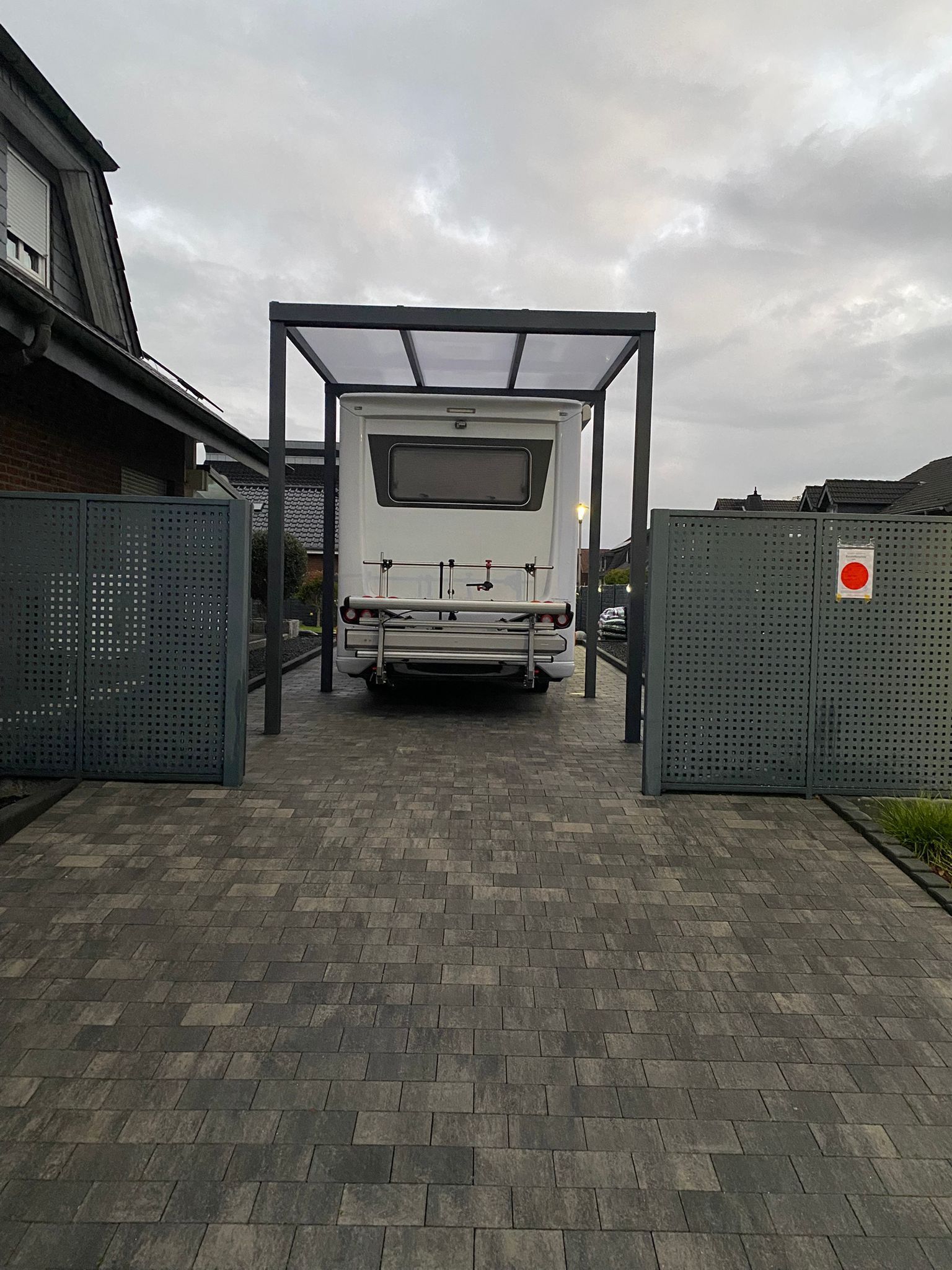 Ein weißes Wohnmobil parkte unter einem modernen, nach einer Seite offenen Metallunterstand auf einer gepflasterten Auffahrt zwischen zwei grauen Sichtschutzwänden aus Metall.