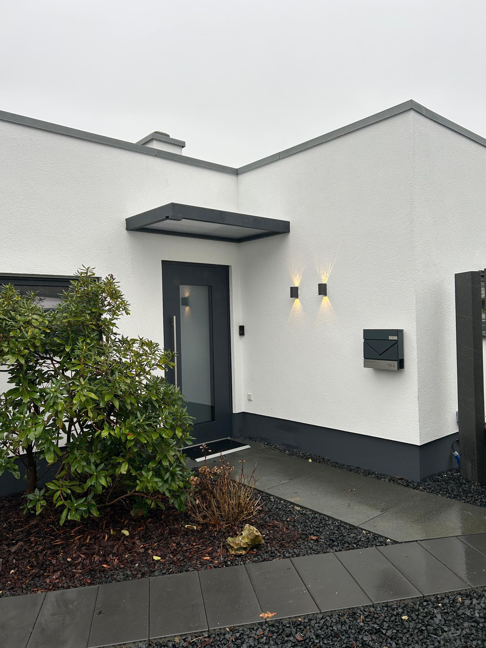 Ein moderner Hauseingang mit weiß strukturierten Wänden, einer dunkelgrauen Tür, einer Markise, Wandleuchten und einem kleinen Garten.