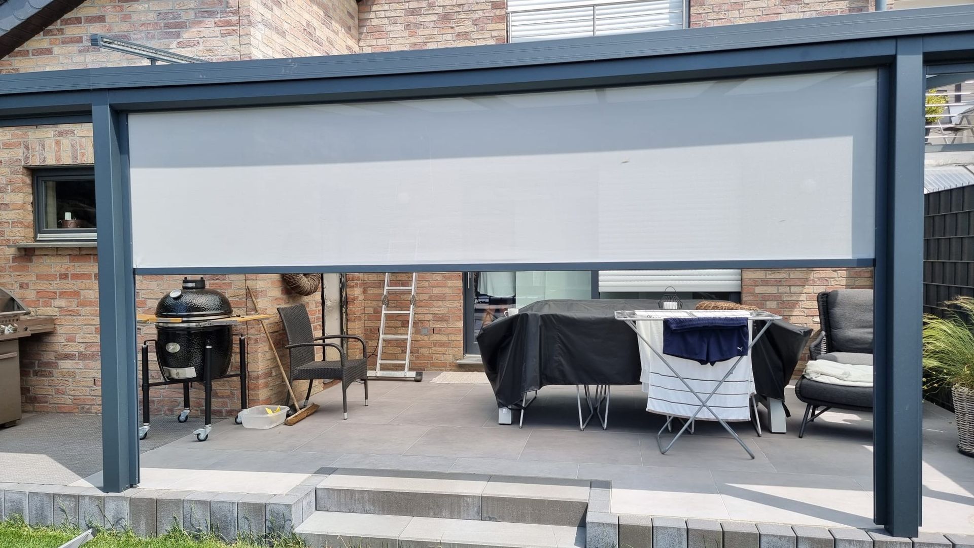 Eine helle, motorisierte Außenwand für die Terrasse, die teilweise unter einem dunklen Rahmen auf einer mit Ziegeln ummauerten Terrasse heruntergelassen ist.