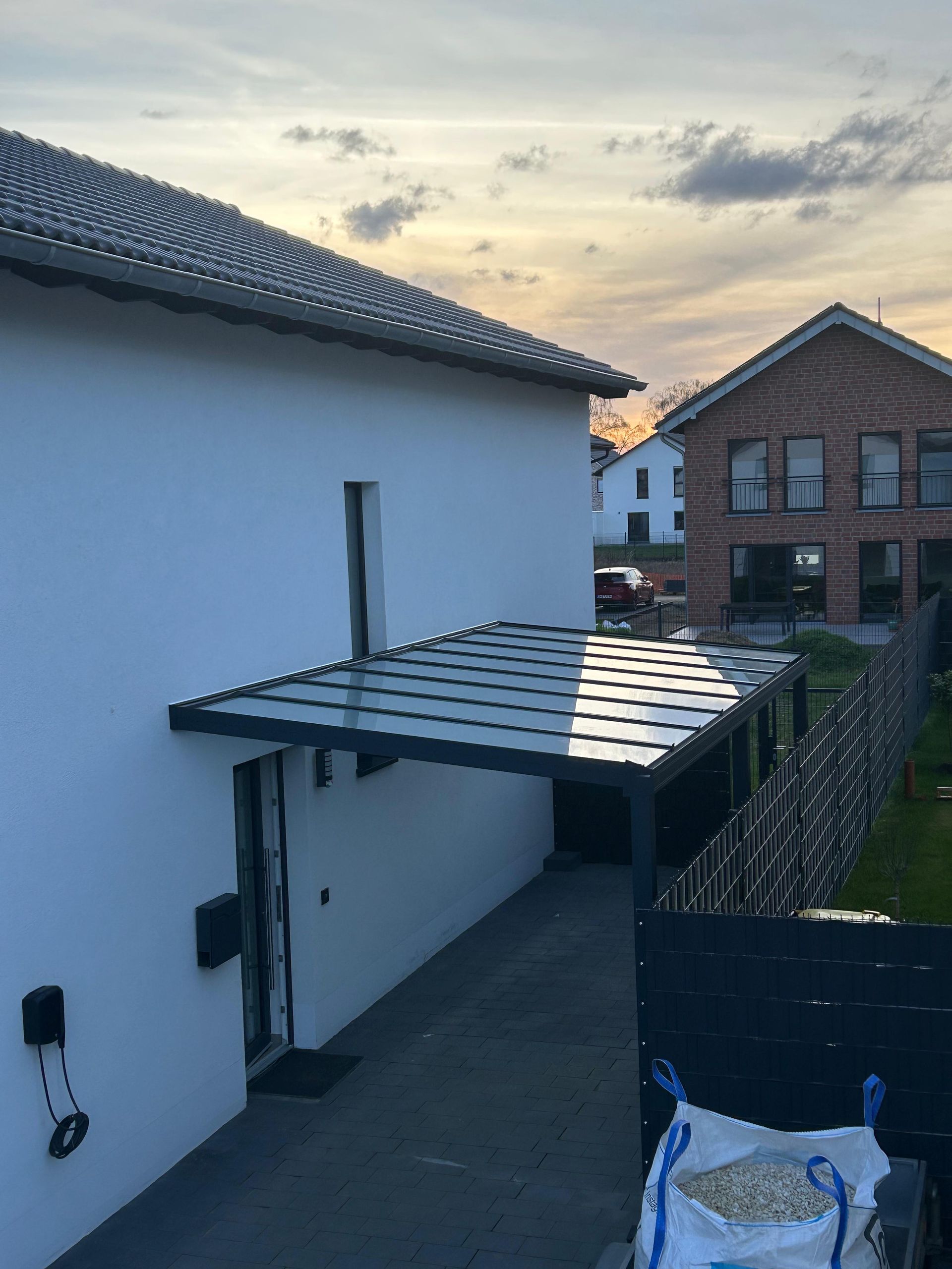 Ein modernes weißes Haus mit einer dunklen, metallgerahmten Terrassenüberdachung, die sich bei Sonnenuntergang über eine Betonauffahrt erstreckt.