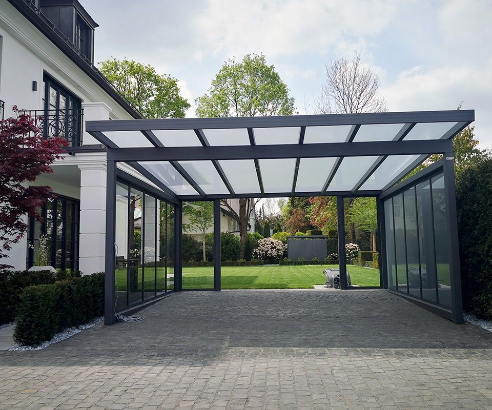 Ein dunkelgrauer Carport aus Metall mit lichtdurchlässigem Dach, der an der Seite eines modernen weißen Hauses mit Steinauffahrt angebaut ist.