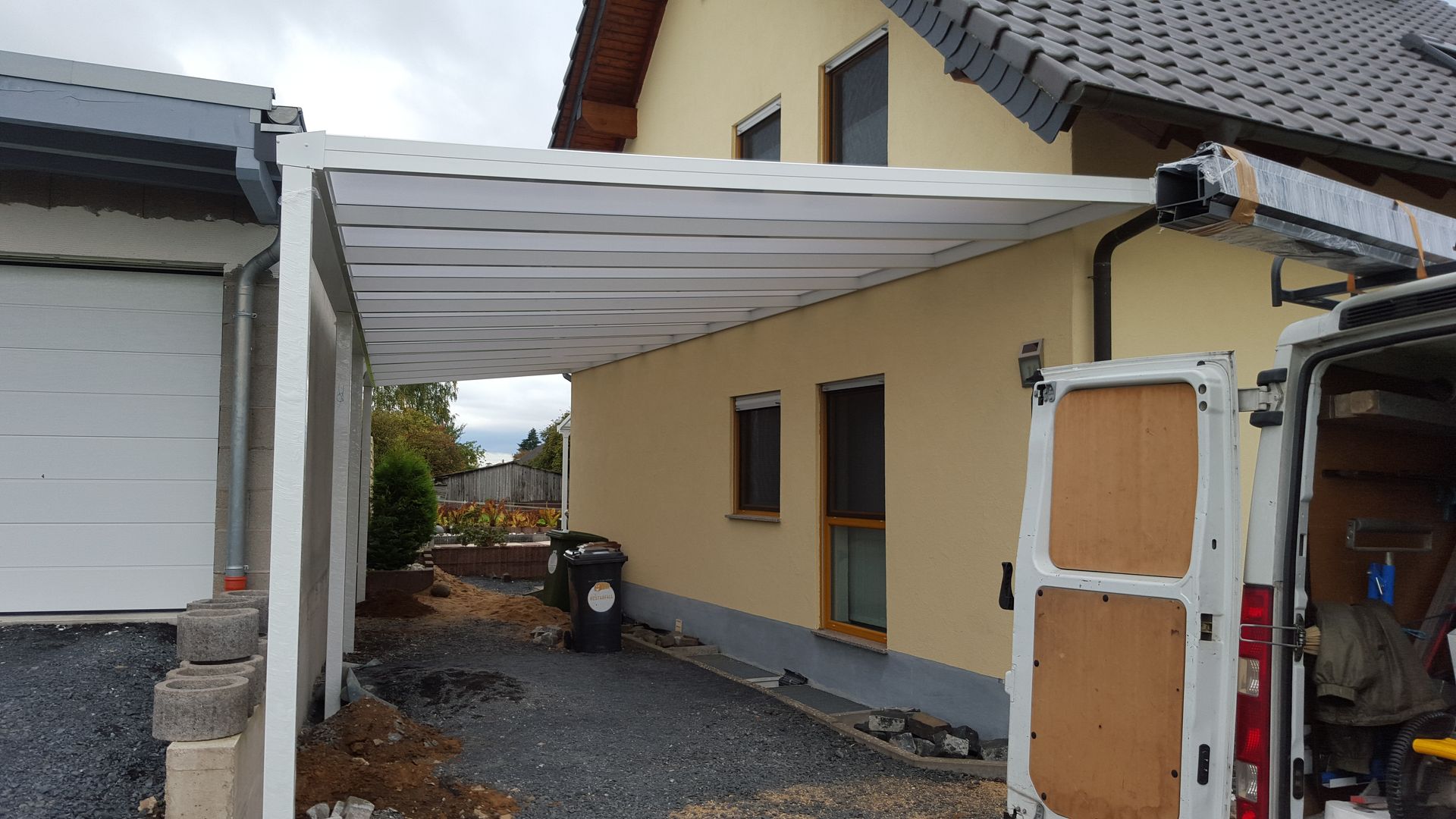 Ein weißes Carport oder eine Terrassenüberdachung ist an ein gelbes Haus angebaut, im Vordergrund ist ein Lieferwagen geparkt.