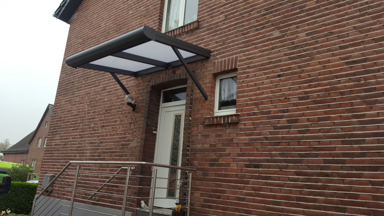 Die Fassade eines Hauses aus roten Backsteinen weist eine moderne Eingangstür mit Metallgeländer und ein flaches, schwarz umrahmtes Vordach darüber auf.