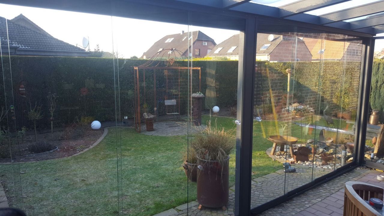 Blick von einer überdachten Terrasse durch Glasscheiben in einen grünen Garten mit Gartenschaukel, kleinen Bäumen und einer Terrassenfläche.