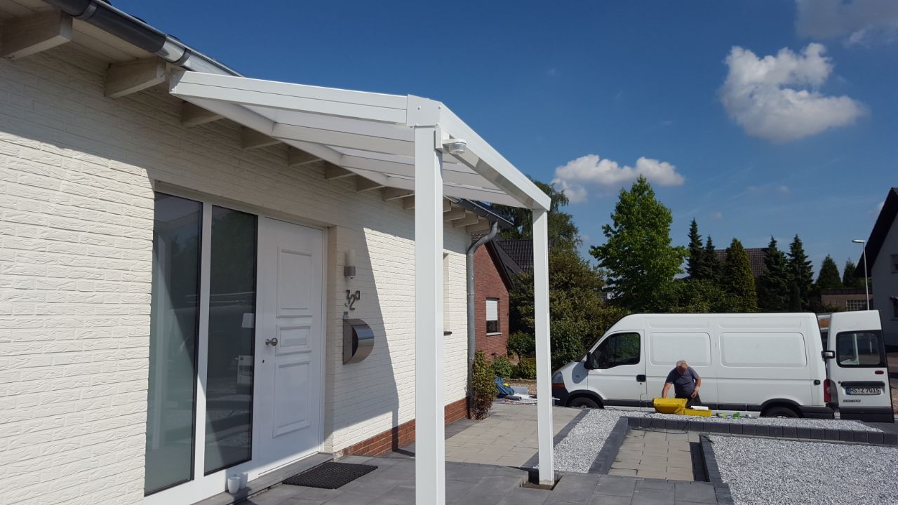 Ein weißes Haus mit modernem, überdachtem Eingangsbereich, einer gepflasterten Auffahrt und im Hintergrund geparkten weißen Lieferwagen.