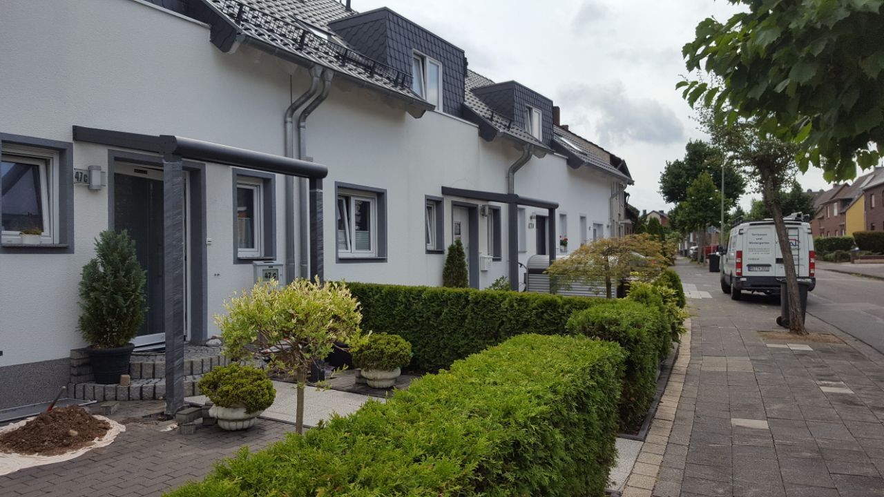 Eine Reihe weißer Stadthäuser mit grauen Zierelementen, kleinen Vorgärten und einem Bürgersteig entlang einer ruhigen Wohnstraße.