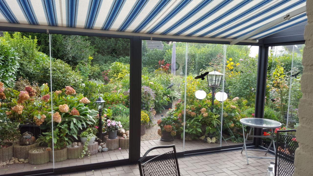 Eine überdachte Terrasse mit blau-weiß gestreifter Markise, Glaswänden und Blick auf einen üppigen, blühenden Garten.