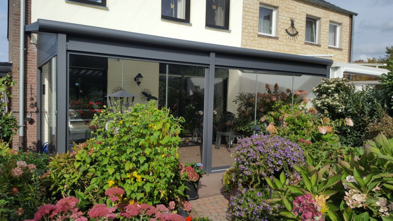 Eine moderne, graue Terrassenüberdachung mit Glaswänden, die an ein Wohnhaus angebaut ist und von einem üppigen, blühenden Garten umgeben ist.
