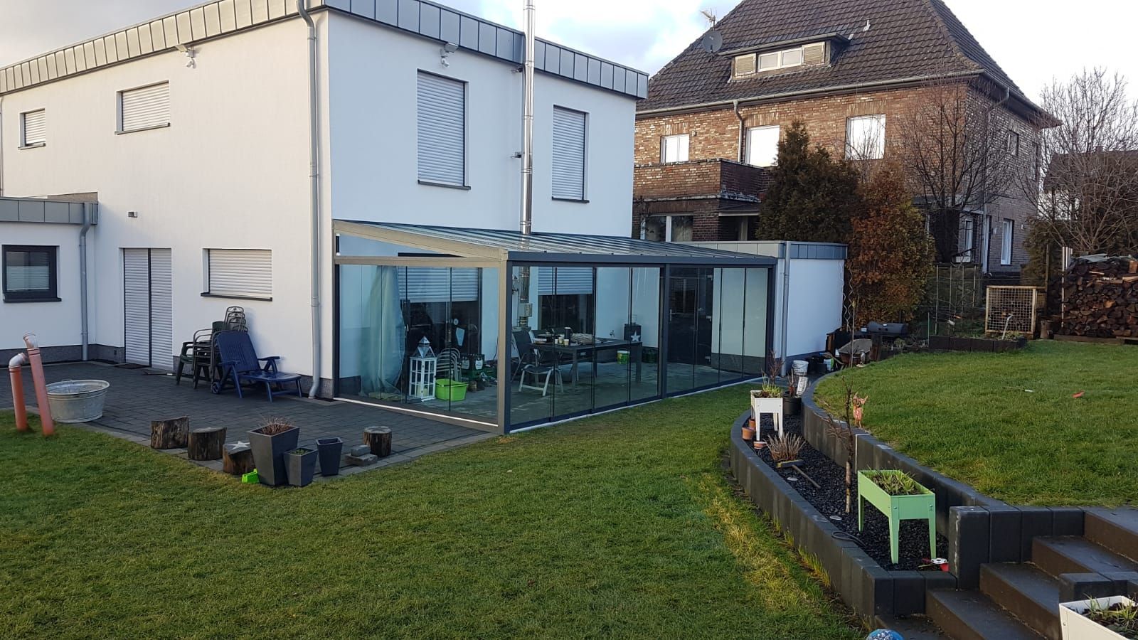 Ein weißes Haus mit einer modernen, verglasten Terrasse, eingebettet in einen Garten mit Rasen, Beeten und einem angrenzenden Backsteinhaus.