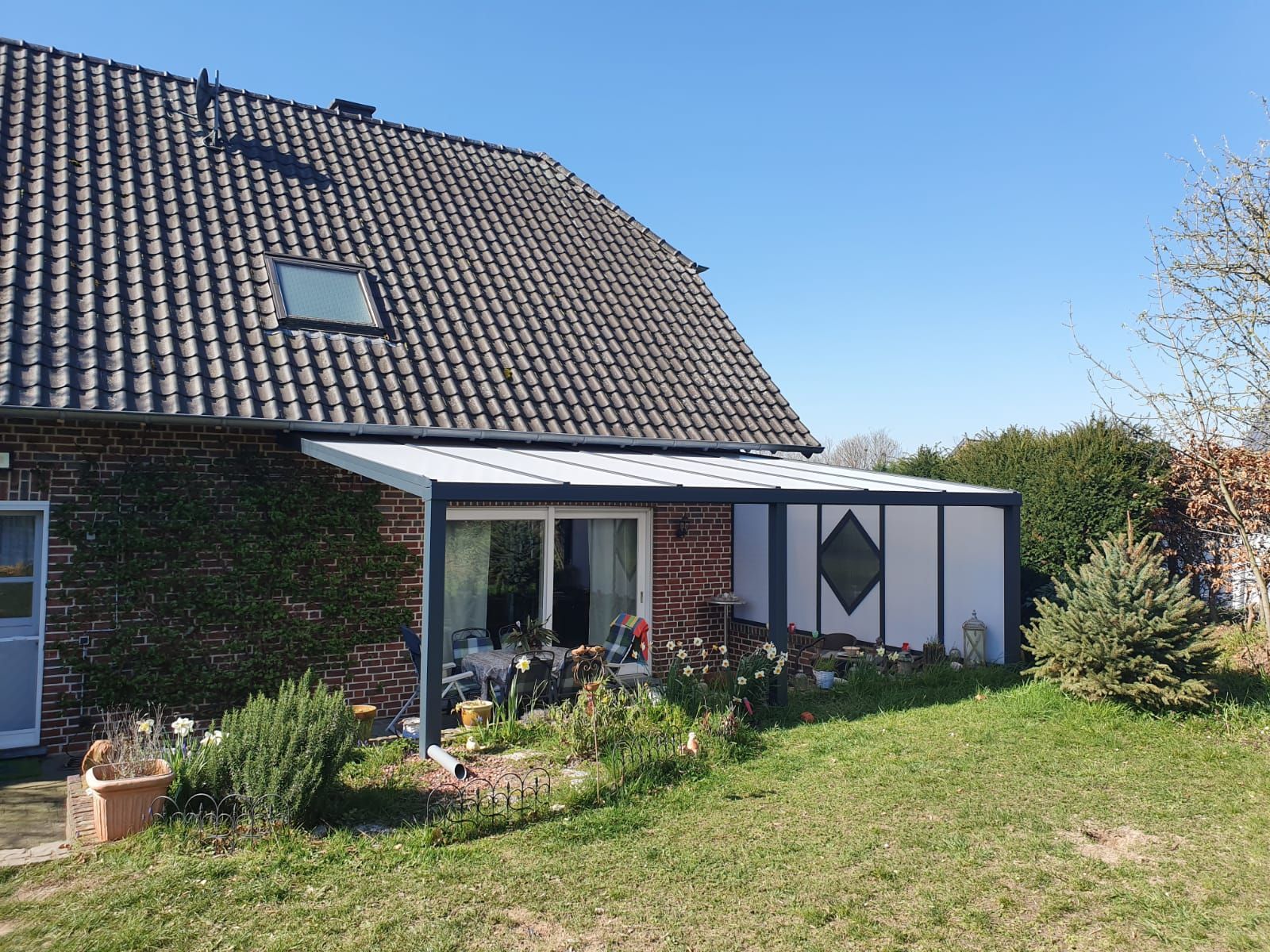 Ein Backsteinhaus mit einer weiß umrahmten Terrassenüberdachung und einem Glasdach, das sich über einen Gartenbereich unter klarem, blauem Himmel erstreckt.