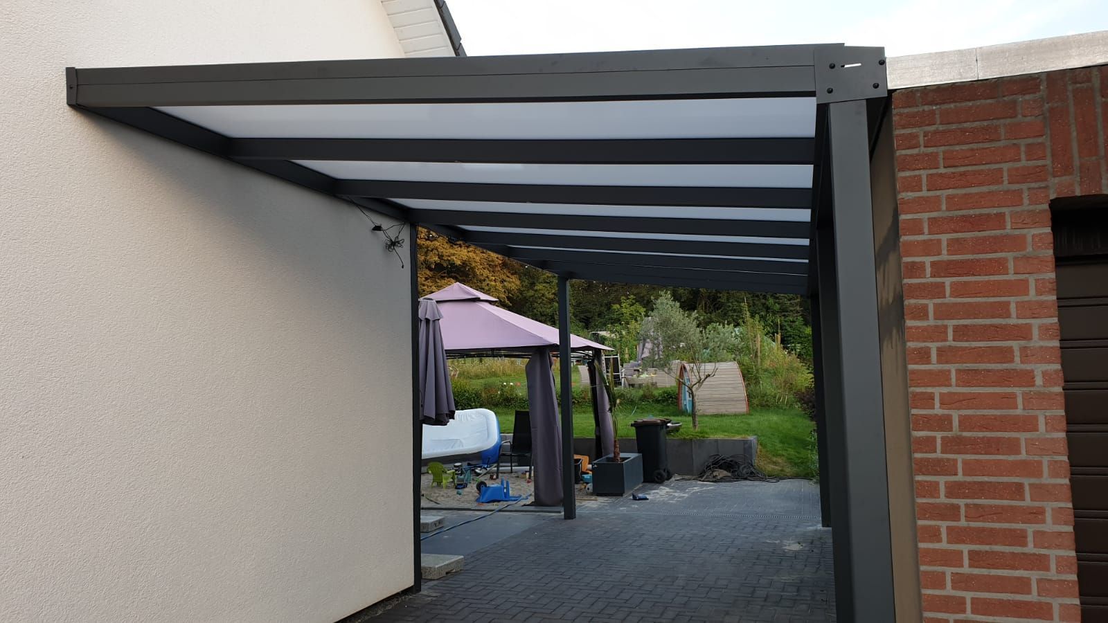 Eine dunkelgraue Terrassenüberdachung mit weißem, lichtdurchlässigem Dach, die an einer beigefarbenen Wand und einem Gebäude aus roten Ziegelsteinen befestigt ist.