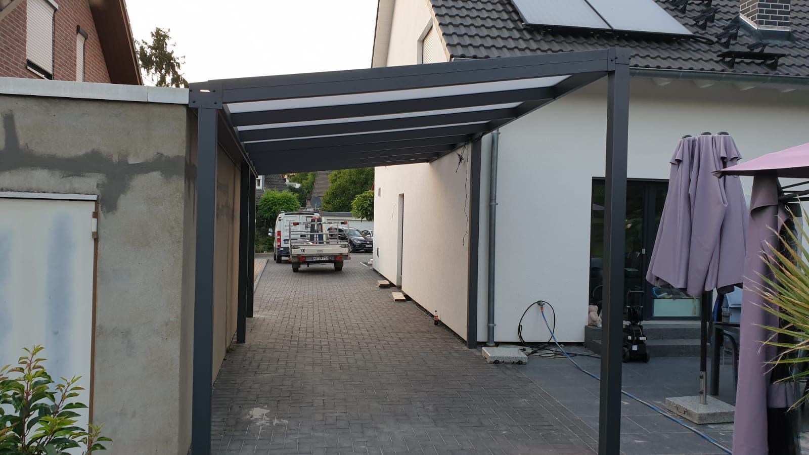 Zwischen einem weißen Haus und einer Betonmauer über einer gepflasterten Auffahrt befindet sich ein moderner, dunkel umrahmter Carport mit transparentem Dach.