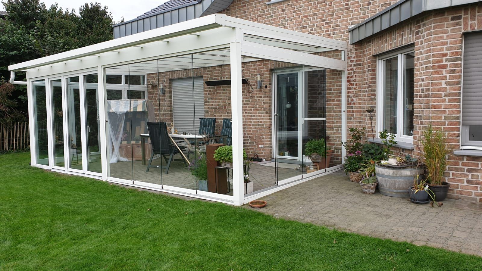 Eine weiß umrahmte Glasterrasse, die an ein Backsteinhaus angebaut ist und einen Blick auf einen grünen Rasen und eine Steinterrasse bietet.