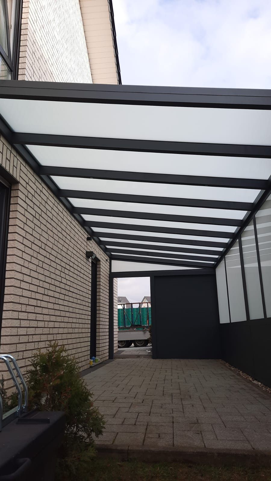 Eine überdachte Terrasse, die an ein Backsteinhaus angrenzt und sich durch ein dunkles Metallgerüst, ein Glasdach und einen gepflasterten Steinboden auszeichnet.